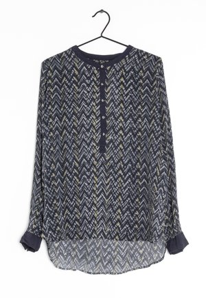 Blouse bleu marine à manches longues avec motif en zigzag gris et jaune, patte de boutonnage et poignets arrondis, suspendue à un cintre noir.