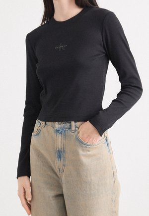 Femme portant un haut noir à manches longues Calvin Klein rentré dans un jean taille haute délavé clair, avec une main dans une poche.