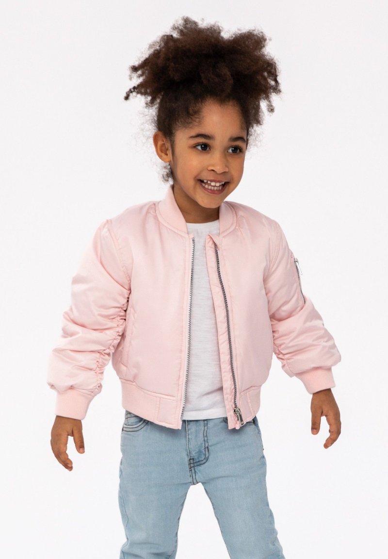 Veste bomber rose en satin, avec des manches froncées, une fermeture éclair à l'avant et des poignets côtelés, associée à un jean bleu clair et un top blanc.