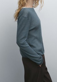 Pull en bleu sarcelle foncé, en tissu texturé. Col rond, manches longues, coupe décontractée. Associé à un pantalon marron, vue de côté contre un fond neutre.