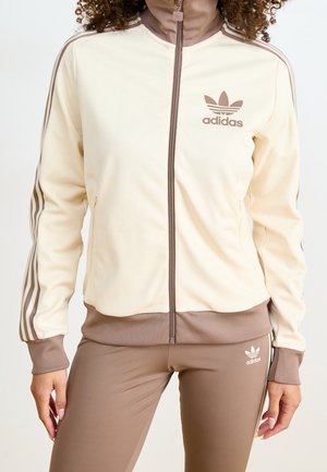 Frau trägt beigen und braunen Adidas-Trainingsanzug mit Reißverschlussjacke und passender Hose, versehen mit Adidas-Logo und drei Streifen an Ärmeln und Beinen.