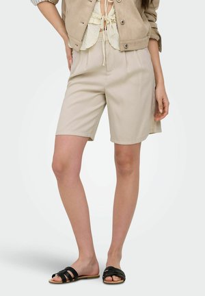 Kvinne iført beige høytlivs knelange shorts, kremfarget cropped topp, beige jakke og svarte sandaler med åpen hæl, stående med hendene langs siden.