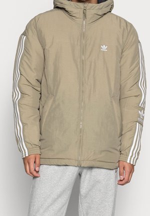 Homme portant une veste à capuche beige Adidas avec des bandes blanches sur les manches et un logo sur la poitrine et le bras, assortie à un pantalon de survêtement gris clair.