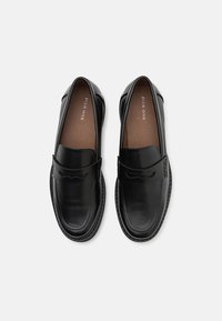 Schwarze Lederloafers mit abgerundetem Zehenbereich, ausgestattet mit einem dekorativen Riemen und Stitching-Details. Das Innere ist braun, was den Kontrast verstärkt.