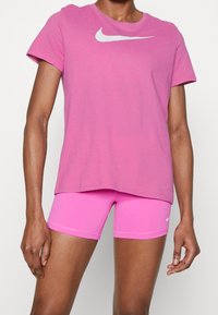 Růžové tričko Nike s krátkým rukávem a bílým logem, v kombinaci s odpovídajícími růžovými sportovními šortkami, které mají hladkou texturu.