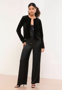 Černé sametové sako s kulatým výstřihem a předními kapsami, vrstvené přes černý saténový top a široké kalhoty černé barvy, s páskovými podpatky.