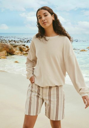 Młoda kobieta o długich kręconych włosach, ubrana w beżową bluzę i pasiasta krótkie spodenki, stoi na piaszczystej plaży z skałami i oceanem w tle.