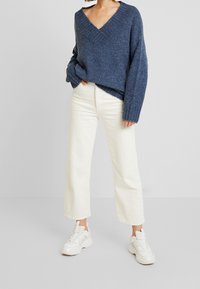 Maglione lavorato a maglia blu navy con scollo a V ampio, abbinato a pantaloni bianchi a gamba larga e sneaker bianche. Design testurizzato e dal taglio rilassato.