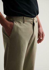 Khaki bukser med elastisk talje, forlommer og lynlås. Glat tekstur med en skræddersyet fit, vist sammen med en sort skjorte.