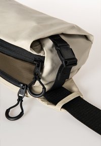 Borsa beige a chiusura arrotolata con una cerniera nera, tracolla piatta nera e dettagli in metallo neri, realizzata in un materiale testurizzato e resistente all'acqua.