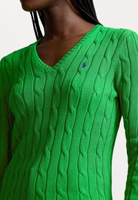 Suéter verde de punto con cuello en V, que presenta patrones de trenzado y un pequeño logotipo azul de acento. El material tiene un acabado texturizado.