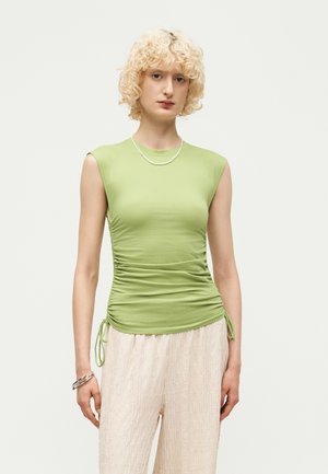 Personne aux cheveux blonds courts et bouclés portant un haut vert clair sans manches froncé et un pantalon ample crème texturé sur un fond uni.