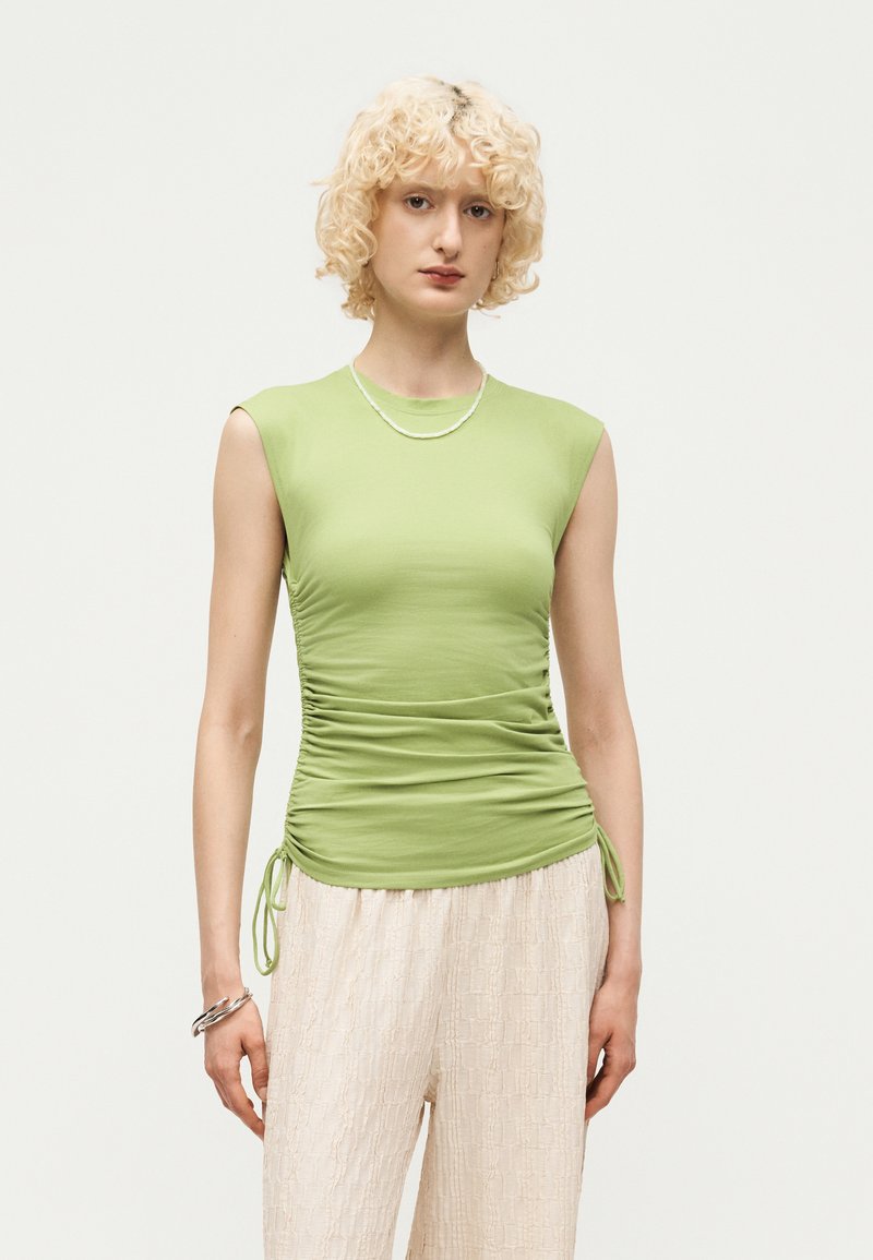 Άτομο με κοντά σγουρά ξανθά μαλλιά, που φορά αμάνικο ανοιχτό πράσινο τοπ με ρυτίδες και κρεμ υφασμάτινο φαρδύ παντελόνι σε μονόχρωμο φόντο.