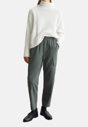 Osoba nosząca biały dziergany sweter z golfem, zielone spodnie typu cargo z boczną kieszenią, czarne skarpetki i czarne buty wsuwane.