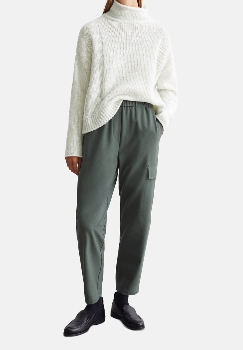 Personne portant un pull en maille blanche à col roulé, un pantalon cargo vert avec poche latérale, des chaussettes noires et des chaussures noires sans lacets.