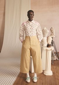 Femeie purtând o bluză cu flori, pantaloni bej cu picioare largi și adidași albi cu șireturi turcoaz, stând lângă busturi sculptate clasice pe piedestale.