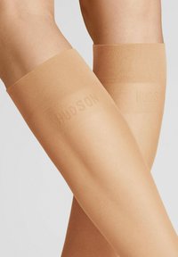 Beige Overknee-Strümpfe mit einer weichen Textur, ausgestattet mit einem glatten Bund oben, der mit dem Markennamen "HUDSON" geprägt ist.