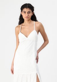 Rochie albă fără sleve, cu decolteu în V, având un model texturat și o fante pe lateral. Fabricată dintr-un material ușor, cu o croială lejeră.