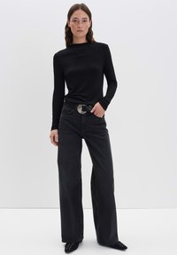 Frau steht gegen eine schlichte Wand, trägt ein schwarzes Langarmoberteil, weite schwarze Jeans mit Gürtel und schwarze spitze Schuhe.