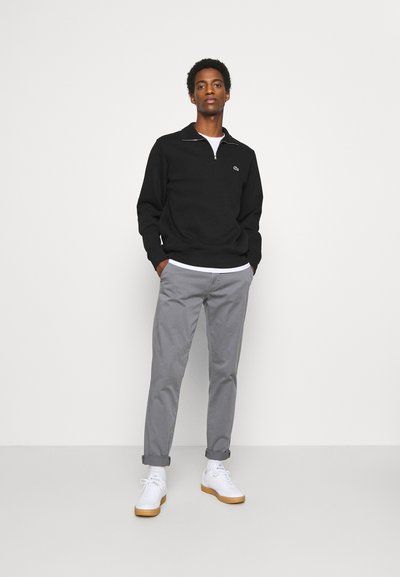 Sweatshirt noir zippé avec des accents blancs, pantalon droit gris et baskets blanches avec des semelles en caoutchouc. Tenue simple et décontractée sur un fond uni.