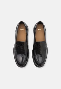 Mocassins en cuir noir avec une finition brillante, arborant un détail de logo en relief à l'avant, une texture lisse et une semelle intérieure rembourrée.