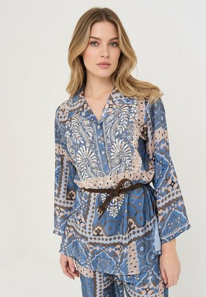 Femme blonde portant un chemisier et un pantalon à motifs floraux et géométriques bleu, beige et marron, avec une ceinture noire nouée à la taille.