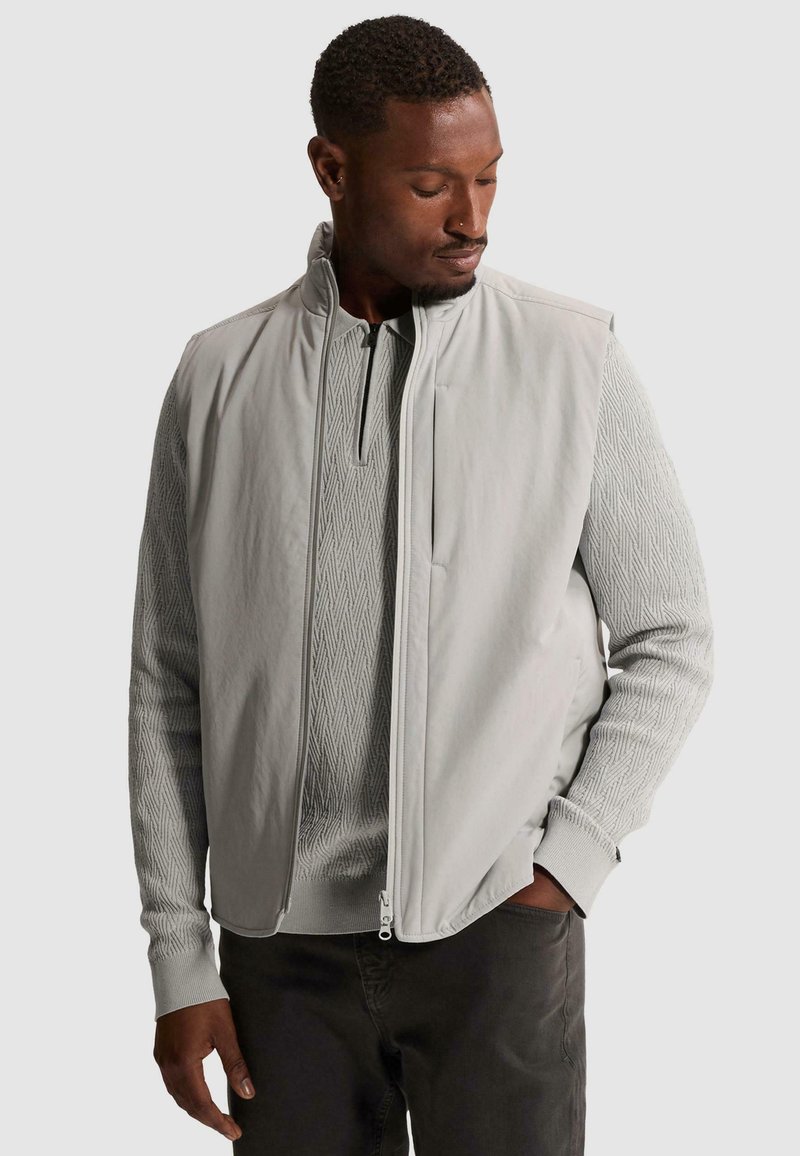 Homme portant un gilet matelassé gris clair sur un pull à manches longues à motifs, regardant vers le bas avec une main dans la poche d'un pantalon sombre.