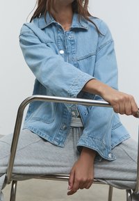 Une personne dans une veste en denim bleu clair et un pantalon de survêtement gris est assise sur une chaise en métal, tenant l'accoudoir de la chaise avec une main.