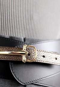 Curea din piele cu o bandă texturată de bronz metalic și un cataramă aurie lucioasă. Are un suport negru și găuri vizibile pentru curea.