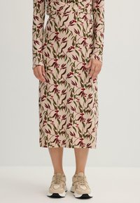 Falda midi de tela beige con patrón floral en rojo y verde; cintura ajustada, mangas largas, combinada con zapatillas ligeras beige.