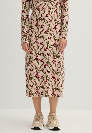 Falda midi de tela beige con patrón floral en rojo y verde; cintura ajustada, mangas largas, combinada con zapatillas ligeras beige.