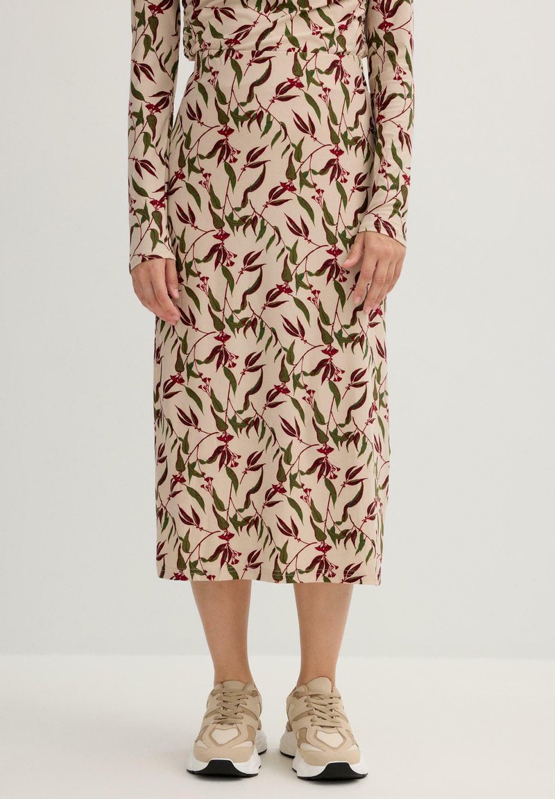 Falda midi de tela beige con patrón floral en rojo y verde; cintura ajustada, mangas largas, combinada con zapatillas ligeras beige.