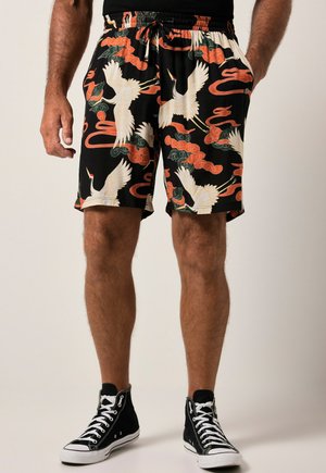 Man draagt zwarte shorts met witte kraanvogels en oranje wolkenpatronen, gecombineerd met zwarte hoge sneakers en zwarte sokken.