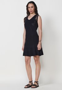 Schwarzes ärmelloses Kleid mit tailliertem Oberteil, ausgestellt am Saum, gerippter Textur und floralen Stickereidetails nahe der Schulter. Getragen mit Riemchensandalen.