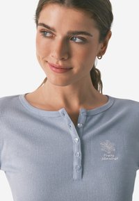 Ung kvinde med brunt hår trukket tilbage, iført en lys blå ribbet skjorte med knapper og en lille snefnug brodering på brystet.