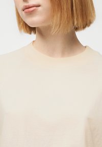 Ανοιχτό μπεζ φούτερ με ριμπ λαιμόκοψη και λεία υφή. Το ύφασμα πέφτει απαλά, με διακριτικές ραφές στους ώμους.