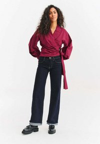 Bordowa bluzka kopertowa z bufkami, zestawiona z ciemnoniebieskimi szerokimi dżinsami i czarnymi butami na platformie z ozdobami w formie frędzli.