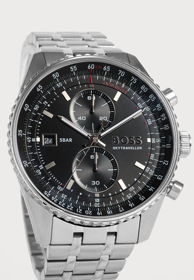 Montre-bracelet en métal argenté avec cadran noir, tachymètre, deux sous-cadrans, affichage de la date et marque "BOSS Skytraveller", affichant l'heure 10:09:40.