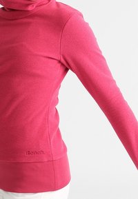 Top de forro polar rosa, ajustado y de manga larga, con un alto cuello y un dobladillo acanalado. Presenta un logotipo sutil cerca del dobladillo y una textura lisa.