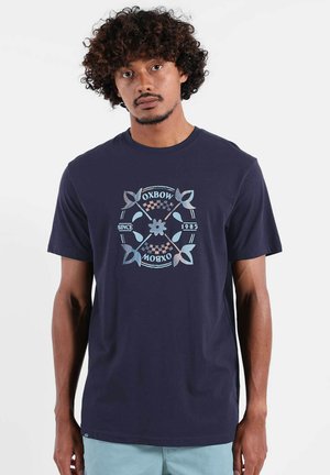 Homme aux cheveux bouclés portant un t-shirt Oxbow bleu marine avec un motif floral et à carreaux, associé à un short bleu clair, debout devant un fond blanc.