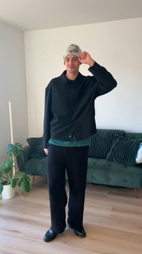 Giacca nera con bottoni sopra una maglia verde, abbinata a pantaloni neri e scarpe nere. Un berretto chiaro completa l'outfit.