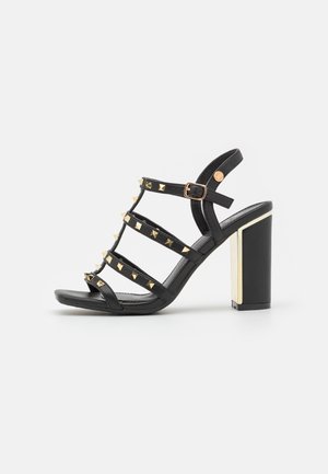 Sandalia negra con múltiples tiras y tachuelas doradas, tacón grueso con un acento dorado, fabricada en material sintético, acabado mate.