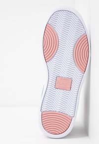 Bílé tenisky s texturovanou gumovou podrážkou, na které jsou růžové kruhové akcenty. Podešev má vlnitý vzor a viditelnou logo nášivku.