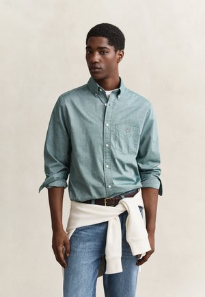 Junger Mann trägt ein grünes Hemd mit Knöpfen und hochgekrempelten Ärmeln, blaue Jeans und einen weißen Pullover, der um die Taille gebunden ist, vor einem einfarbigen Hintergrund.