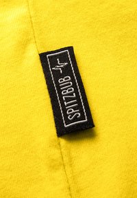 Gelbes Baumwollgewebe mit einer strukturierten Oberfläche, das mit einem schwarzen Stoffetikett versehen ist, auf dem in weißer Schrift "SPITZBUB" steht, das am Rand genäht ist.