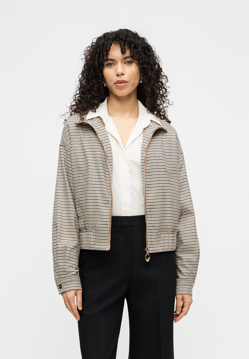 Καρό μπουφάν με φερμουάρ σε μπεζ και μαύρο, με γιακά όρθιο, ελαστικά μανίκια και χρυσή λεπτομέρεια στο φερμουάρ, σε συνδυασμό με λευκό πουκάμισο και μαύρο παντελόνι.