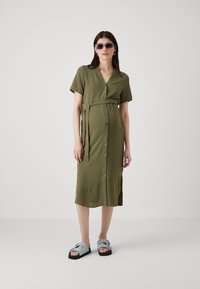 Olivenfarget, kortermet kjole med V-hals, knappefront, belte i livet og sidesplitte. Modellen har på seg lyseblå sandaler og solbriller.