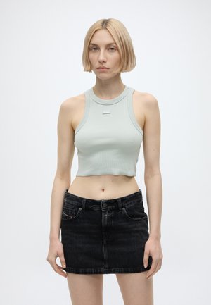 Jeune femme aux cheveux blonds courts portant un crop top gris clair sans manches et une mini-jupe en jean noire sur un fond uni.