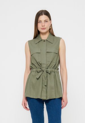 Naine kannab varrukateta oliivrohelise vöökohaga särki, millel on krae ja ees taskud, koos siniste teksadega, seisab valge taustaga.