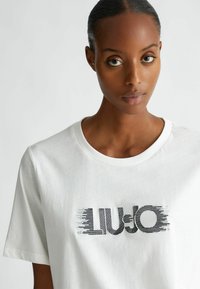 T-shirt di cotone bianco con scollatura rotonda e maniche corte. Caratterizzato da un logo grafico nero "LIU JO" con elementi di design lineare.